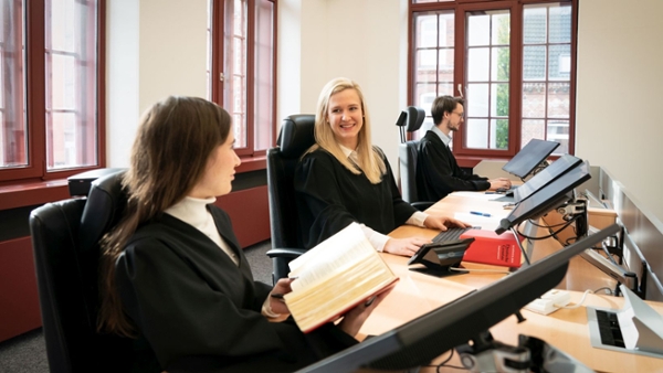 Drei Menschen sitzen in einem Gerichtssaal. (Quelle: Staatskanzlei) Drei Menschen sitzen in einem Gerichtssaal.